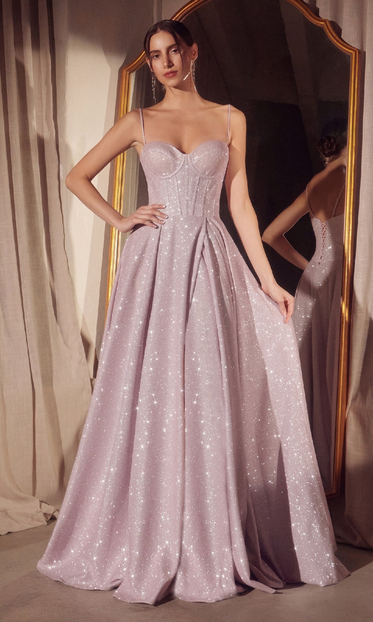 A woman stands in front of a mirror, holding one side of her Lace-Up Long Glitter Prom Ball Gown: Amoris T706, a sparkling light lavender A-line evening dress with thin straps.